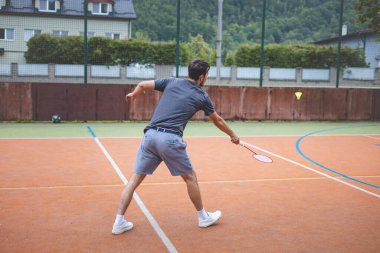 Badminton oyuncusu açık hava sahasında oynanan bir maçta havadaki pipiye odaklanıyor. Raketi yükselirken, kentsel ve manzaralı bir arka planla çevrili bir sonraki çekim için hazırlanıyor..