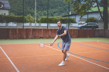 Badminton oyuncusu açık hava sahasında oynanan bir maçta havadaki pipiye odaklanıyor. Raketi yükselirken, kentsel ve manzaralı bir arka planla çevrili bir sonraki çekim için hazırlanıyor..