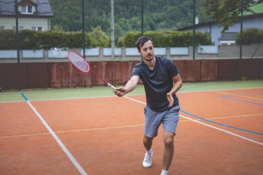 Badminton oyuncusu açık hava sahasında oynanan bir maçta havadaki pipiye odaklanıyor. Raketi yükselirken, kentsel ve manzaralı bir arka planla çevrili bir sonraki çekim için hazırlanıyor..