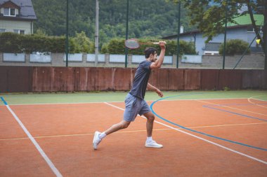 Badminton oyuncusu açık hava sahasında oynanan bir maçta havadaki pipiye odaklanıyor. Raketi yükselirken, kentsel ve manzaralı bir arka planla çevrili bir sonraki çekim için hazırlanıyor..