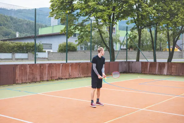 stock image Young man prepares to play badminton on an outdoor court. Holding a racket and shuttlecock, he stands ready to serve in a casual setting surrounded by trees and urban elements.