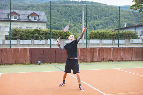 stock image Badminton player stands ready to serve on an outdoor court, holding a racket and shuttlecock. Dressed in casual sportswear, he focuses on the game amid a scenic, natural setting.