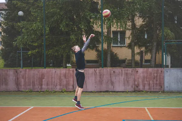 Basketbolcu topu dışarıda bir sahada atmaya hazırlanıyor ve kendini odaklanmış bir atış için hazırlıyor. Sahne, ağaçların ve kentsel binaların arka planıyla atleti hareketin ortasında yakalıyor..