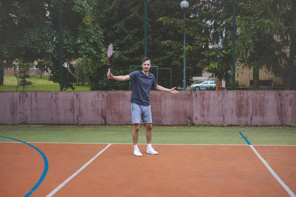 stock image Badminton player focuses on the shuttlecock mid-air during a match on an outdoor court. With his racket raised, he prepares for the next shot, surrounded by an urban and scenic background.