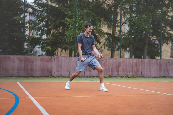 stock image Badminton player celebrates a moment on an outdoor court, holding his racket in an energetic stance. His expression shows excitement and triumph, set against a backdrop of trees and buildings.