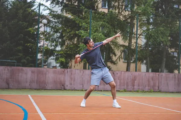 stock image Badminton player celebrates a moment on an outdoor court, holding his racket in an energetic stance. His expression shows excitement and triumph, set against a backdrop of trees and buildings.