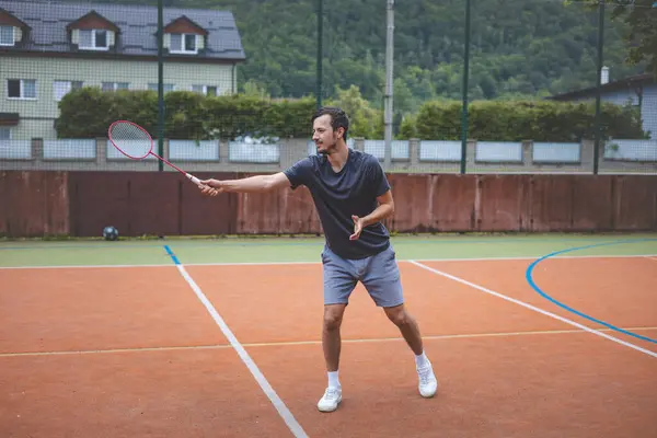 stock image Badminton player focuses on the shuttlecock mid-air during a match on an outdoor court. With his racket raised, he prepares for the next shot, surrounded by an urban and scenic background.