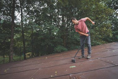 Adam eğimli bir çatıyı ağaçlarla çevrili uzun bir süpürgeyle temizliyor. Açık hava düzenlemesi yemyeşil, şahıs çatıdaki enkazı kaldırma görevine odaklanmış durumda..