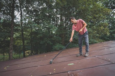 Man is cleaning a slanted roof with a long broom, surrounded by trees. The outdoor setting features lush greenery, with the individual focused on his task of removing debris from the roof. clipart