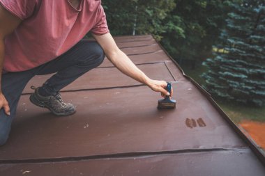 Man crouches down, carefully painting the surface of a roof with a small brush. His focus is on applying even strokes of paint. The outdoor setting is surrounded by trees, creating a peaceful atmosphere. clipart