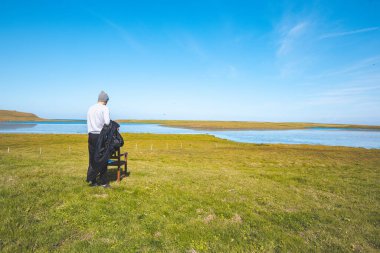 Sıcak bir ceket giymiş bir adam yeşil bir tarlada duruyor, ahşap bir sandalye ayarlıyor, arka planda dingin bir nehir ve mavi gökyüzü, Batı İzlanda 'nın huzurlu manzarasında geçiyor..