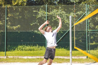 Genç adam havada, güçlü bir voleybol atlama servisini kumlu bir sahada yapıyor, kolunu kaldırıp topa vuruyor. Şiddetli hareket, yeşil bir arka planla çevrili açık havada gerçekleşiyor..