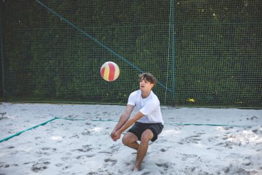 Genç bir adam voleybol pasları atar, topla kumlu bir sahada temasta bulunurken dikkatle odaklanır. Çevresi yeşilliklerle çevrili açık hava onun atletizmini vurguluyor..