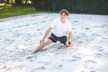 Genç adam, voleybol oynadıktan sonra kumlu bir sahada otururken gülümsüyor. Onun rahat duruşu, yeşilliklerle çevrili dinamik açık hava ortamıyla çelişiyor..