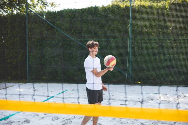 Genç adam kumlu bir kortta dururken bir voleybol topunu şakayla sektiriyor. Maç sırasındaki rahatlama anı spordan zevk aldığını vurguluyor..