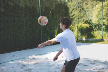 Genç adam kumlu bir kortta dururken bir voleybol topunu şakayla sektiriyor. Maç sırasındaki rahatlama anı spordan zevk aldığını vurguluyor..