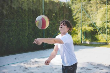 Genç adam kumlu bir kortta dururken bir voleybol topunu şakayla sektiriyor. Maç sırasındaki rahatlama anı spordan zevk aldığını vurguluyor..