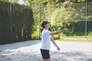 Genç adam kumlu bir kortta dururken bir voleybol topunu şakayla sektiriyor. Maç sırasındaki rahatlama anı spordan zevk aldığını vurguluyor..
