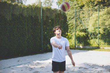 Genç adam kumlu bir kortta dururken bir voleybol topunu şakayla sektiriyor. Maç sırasındaki rahatlama anı spordan zevk aldığını vurguluyor..
