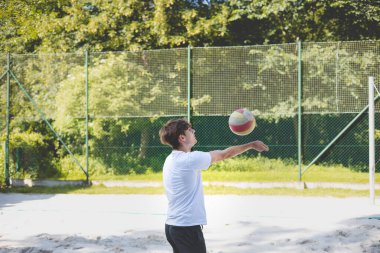 Genç adam kumlu bir kortta dururken bir voleybol topunu şakayla sektiriyor. Maç sırasındaki rahatlama anı spordan zevk aldığını vurguluyor..