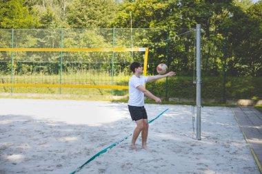 Genç adam kumlu bir kortta dururken bir voleybol topunu şakayla sektiriyor. Maç sırasındaki rahatlama anı spordan zevk aldığını vurguluyor..