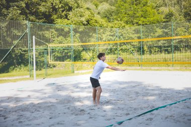 Genç adam kumlu bir kortta dururken bir voleybol topunu şakayla sektiriyor. Maç sırasındaki rahatlama anı spordan zevk aldığını vurguluyor..