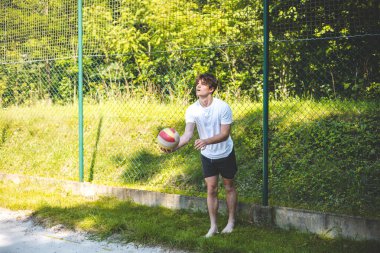 Genç adam bir voleybol maçına hazırlanıyor, elinde odaklanarak kırmızı ve sarı bir top tutuyor. Sahilde sahanın önündeki çimenlikte yalınayak duruyor.