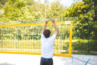 Genç adam plaj voleybolu oynarken voleybolu bloklamak için atlıyor. Odaklanma ve atletizm gösteriyor. Güneşli açık hava sahası yemyeşil manzarayla çevrili..
