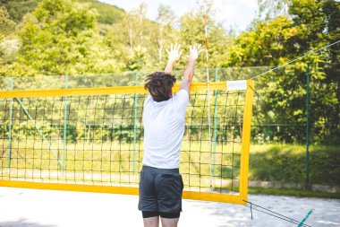 Genç adam plaj voleybolu oynarken voleybolu bloklamak için atlıyor. Odaklanma ve atletizm gösteriyor. Güneşli açık hava sahası yemyeşil manzarayla çevrili..