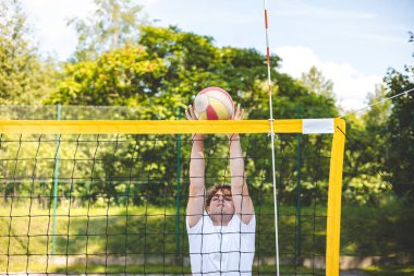 Genç adam plaj voleybolu oynarken voleybolu bloklamak için atlıyor. Odaklanma ve atletizm gösteriyor. Güneşli açık hava sahası yemyeşil manzarayla çevrili..