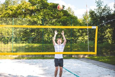 Genç adam plaj voleybolu oynarken voleybolu bloklamak için atlıyor. Odaklanma ve atletizm gösteriyor. Güneşli açık hava sahası yemyeşil manzarayla çevrili..