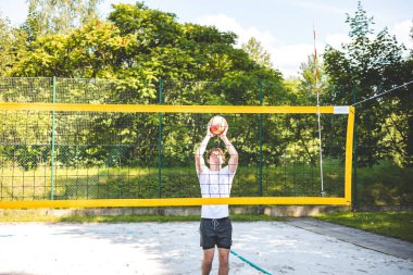 Genç adam plaj voleybolu oynarken voleybolu bloklamak için atlıyor. Odaklanma ve atletizm gösteriyor. Güneşli açık hava sahası yemyeşil manzarayla çevrili..