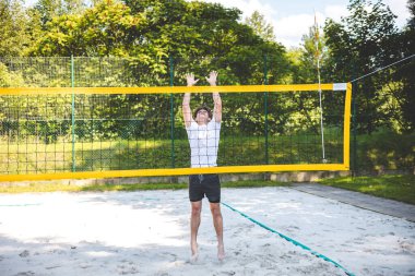 Genç adam plaj voleybolu oynarken voleybolu bloklamak için atlıyor. Odaklanma ve atletizm gösteriyor. Güneşli açık hava sahası yemyeşil manzarayla çevrili..