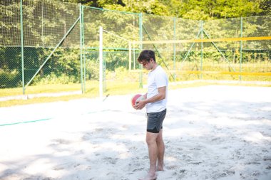 Genç adam bir sonraki oyuna hazırlanmak için kumlu sahadan voleybol topu almak için eğildi. Rahatlamış dış mekan yeşilliklerle çevrili, sıradan bir oyun için mükemmel..