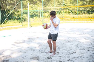 Genç adam bir sonraki oyuna hazırlanmak için kumlu sahadan voleybol topu almak için eğildi. Rahatlamış dış mekan yeşilliklerle çevrili, sıradan bir oyun için mükemmel..