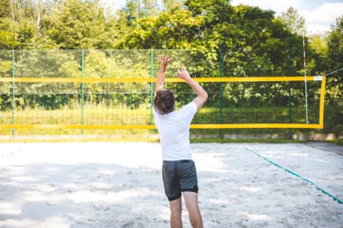 Genç adam atlıyor ve maç sırasında voleybol topuna güçlü bir çivi çakıyor, atletizm ve odaklanmayı gösteriyor. Eylem, yeşilliklerle çevrili güneşli bir kortta gerçekleşiyor..