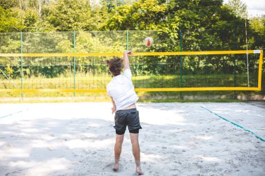 Genç adam atlıyor ve maç sırasında voleybol topuna güçlü bir çivi çakıyor, atletizm ve odaklanmayı gösteriyor. Eylem, yeşilliklerle çevrili güneşli bir kortta gerçekleşiyor..