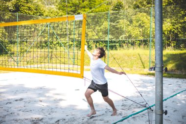 Genç adam atlıyor ve maç sırasında voleybol topuna güçlü bir çivi çakıyor, atletizm ve odaklanmayı gösteriyor. Eylem, yeşilliklerle çevrili güneşli bir kortta gerçekleşiyor..