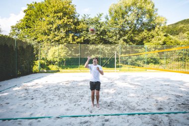 Genç adam, tek eliyle voleybol oynuyor. Açık havada bir antrenman sırasında. Sahne, güneşli, yeşil bir ortamda odak ve beceri gelişimi yakalıyor..