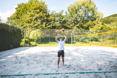 Genç adam, tek eliyle voleybol oynuyor. Açık havada bir antrenman sırasında. Sahne, güneşli, yeşil bir ortamda odak ve beceri gelişimi yakalıyor..