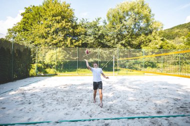 Genç adam, tek eliyle voleybol oynuyor. Açık havada bir antrenman sırasında. Sahne, güneşli, yeşil bir ortamda odak ve beceri gelişimi yakalıyor..