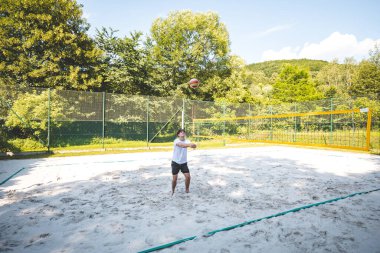 Genç voleybol oyuncusu, kumlu bir sahada ön koluyla pas atarak top alma alıştırması yapıyor. Açık hava mahkemesi yeşillik ve ağaçların arasına kurulmuş doğal bir eğitim ortamı yaratıyor.
