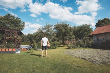 Genç adam bir elinde voleybol topu tutarak yeşil bir arka bahçede yalınayak yürüyor. Bahçe ağaçlarla çevrili ve arka planda kırmızı çatılı, parlak mavi gökyüzünün altında rahat bir ev var..