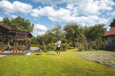 Oyuncu, çim ve ağaçlarla dolu bir bahçede voleybol antrenmanı yapıyor. Beyaz tişört ve siyah şort giymiş, yalınayak. Beyaz bulutlu mavi gökyüzü yaz atmosferini güçlendirir..