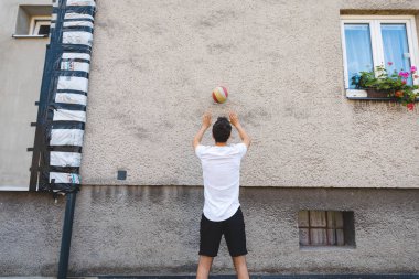 Adam topa sert bir dış duvara vurarak voleybol oynuyor. Duvarın önünde duruyor, beyaz tişört ve siyah şort giyiyor, yeteneklerini geliştirmeye odaklanıyor..