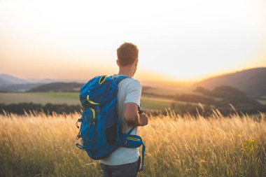 Mavi sırt çantalı bir adam çimenli bir tarlada duruyor, yuvarlanan tepelerin üzerinden nefes kesici bir gün batımını seyrediyor. Sahne, güneş batarken doğanın sükunetini, macerasını ve kişisel yansımasını yakalıyor..