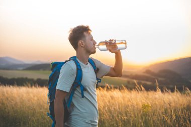 Gün batımında güneşli bir tarlada dikilirken mavi bir sırt çantasıyla gezgin su şişesinden su içer. Altın ışık, uzak tepeler ve barışçıl çevre doğayla olan bağlantıyı vurgular..