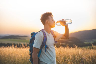Gün batımında güneşli bir tarlada dikilirken mavi bir sırt çantasıyla gezgin su şişesinden su içer. Altın ışık, uzak tepeler ve barışçıl çevre doğayla olan bağlantıyı vurgular..
