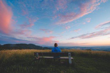 Mavi ceketli bir kadın, uzun otlarla çevrili ve Çek Cumhuriyeti 'nin kayan tepeleri ve vadilerinin manzaralı manzaralı, canlı bir günbatımı gökyüzüne bakan bir bankta oturuyor. Huzurlu ve huzurlu.