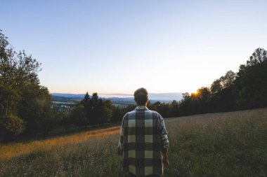 Man in a plaid shirt standing in a grassy meadow, gazing towards the sunset. The sun is low on the horizon, casting a warm glow over the scenic countryside landscape in the Czech Republic. clipart